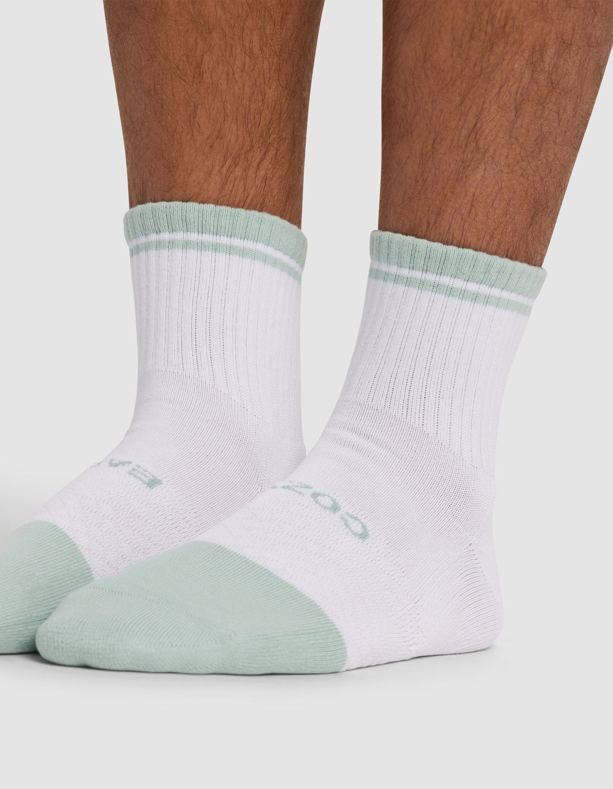 Close-up of a person wearing Cozy Earth’s Essential Quarter Sock - Last Chance in white with light green cuffs, toes, and heels, plus ribbed details and a subtle green stripe near the top, standing on a plain surface.