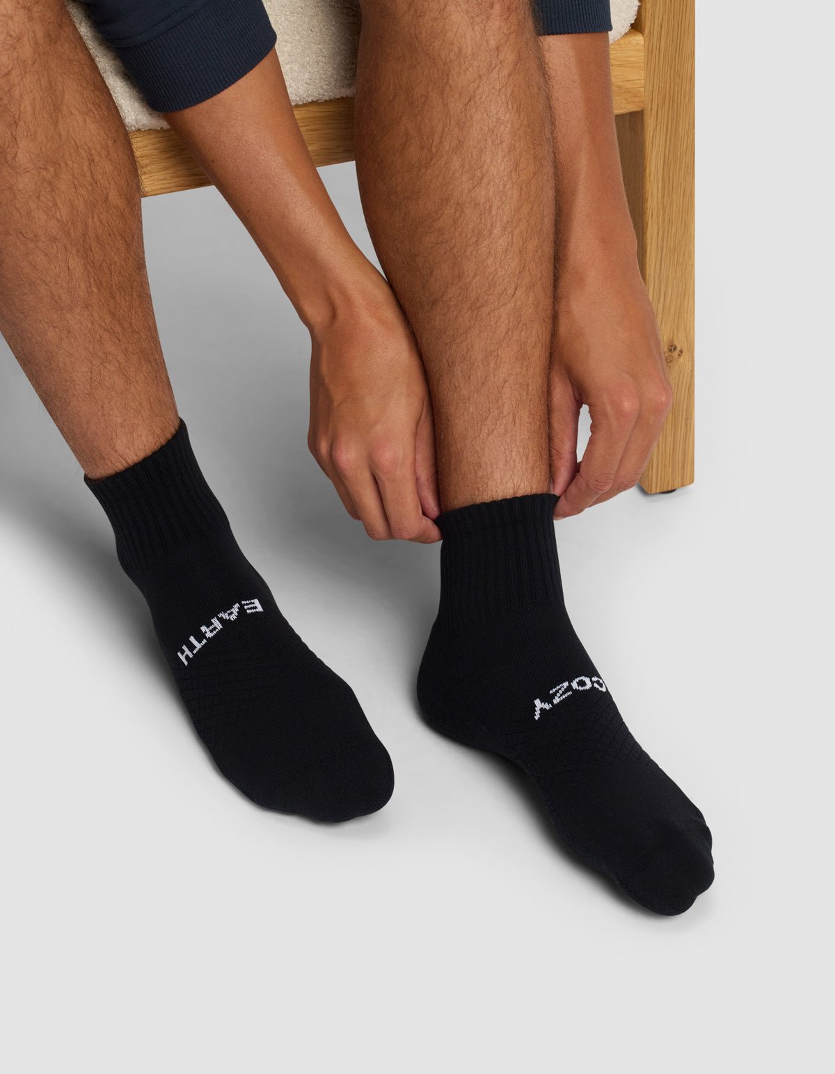 Seated on a wooden chair, a person wearing Cozy Earth Essential Quarter Socks with "EARTH" on the left and "SOCKS" on the right adjusts the right sock with both hands.