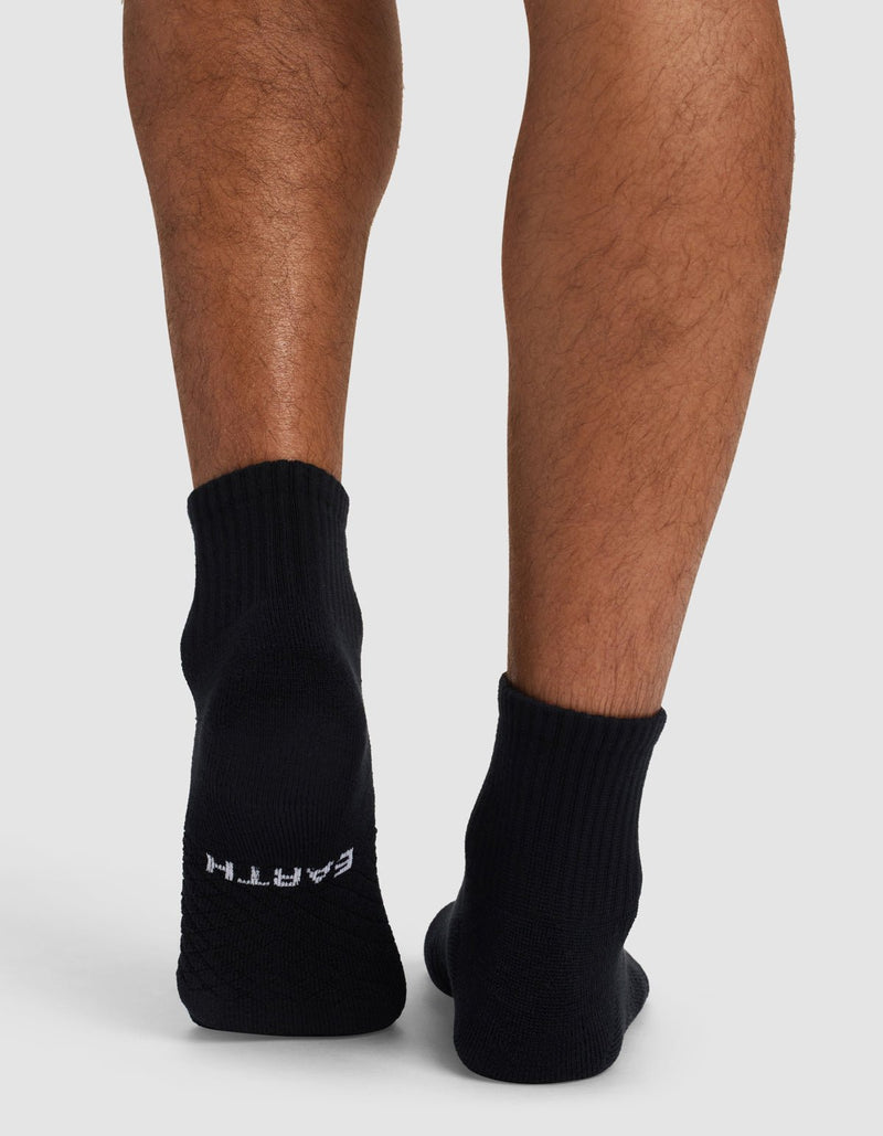 A person’s lower legs and feet are shown from behind, wearing Cozy Earth Essential Quarter Socks in black. The right sock displays "EARTH" in white on the sole. The background is plain gray.