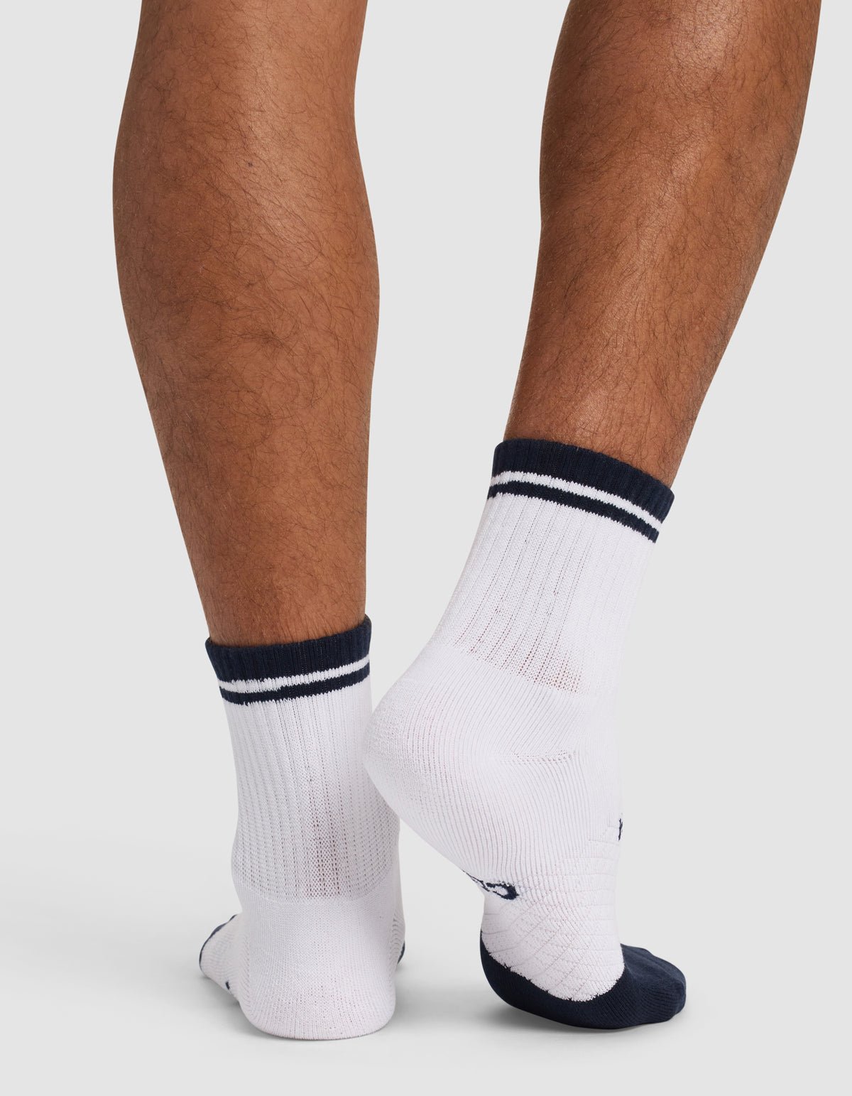 A person stands with crossed feet, shown from the back against a plain white background, wearing Cozy Earth’s Essential Quarter Sock - Last Chance in white with dark blue accents and stripes. 