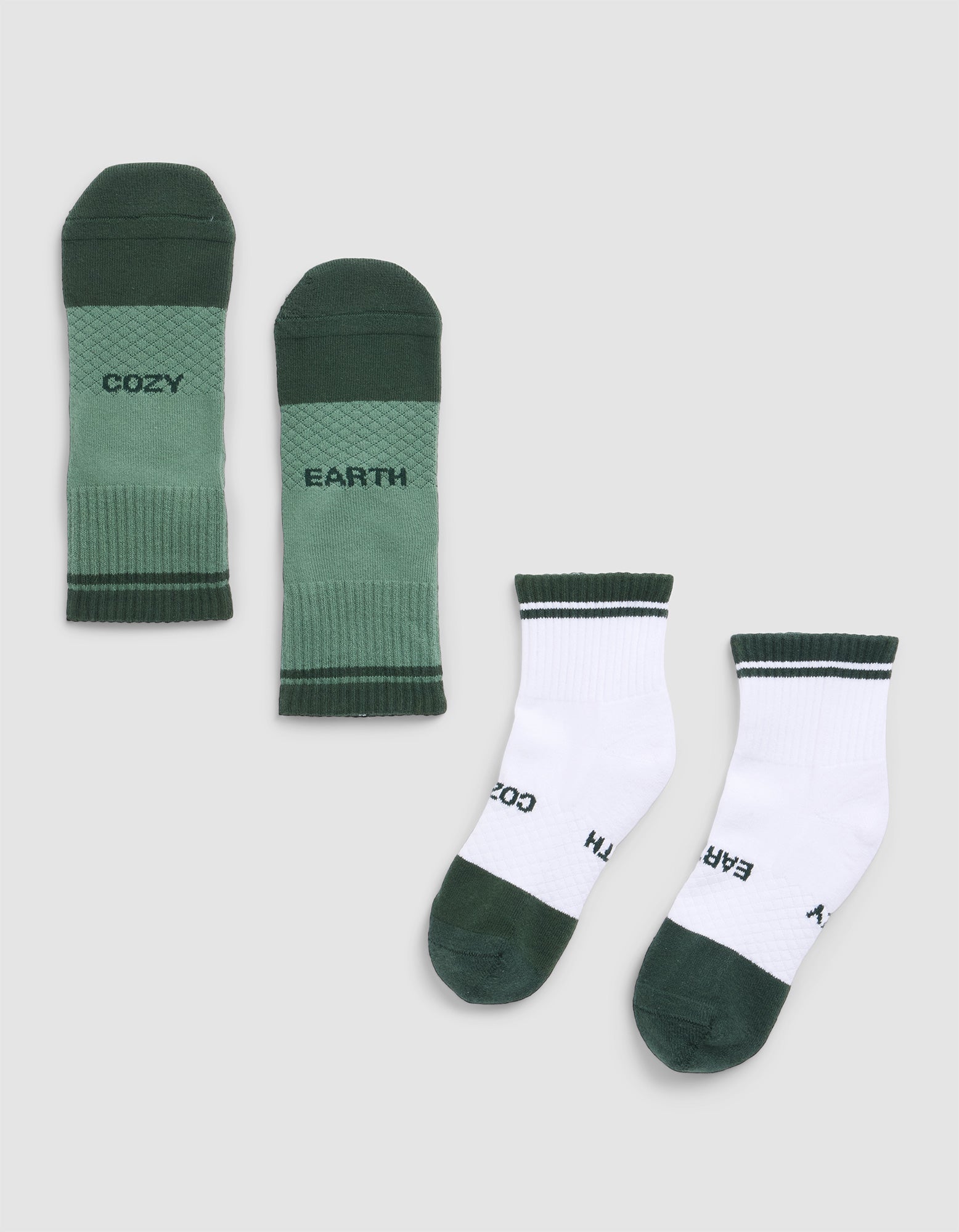 Two pairs of Cozy Earth Essential Quarter Socks are laid flat on a gray background: one pair is green with "COZY" and "EARTH," while the other is white with green toes, heels, and cuffs, featuring the same words. 