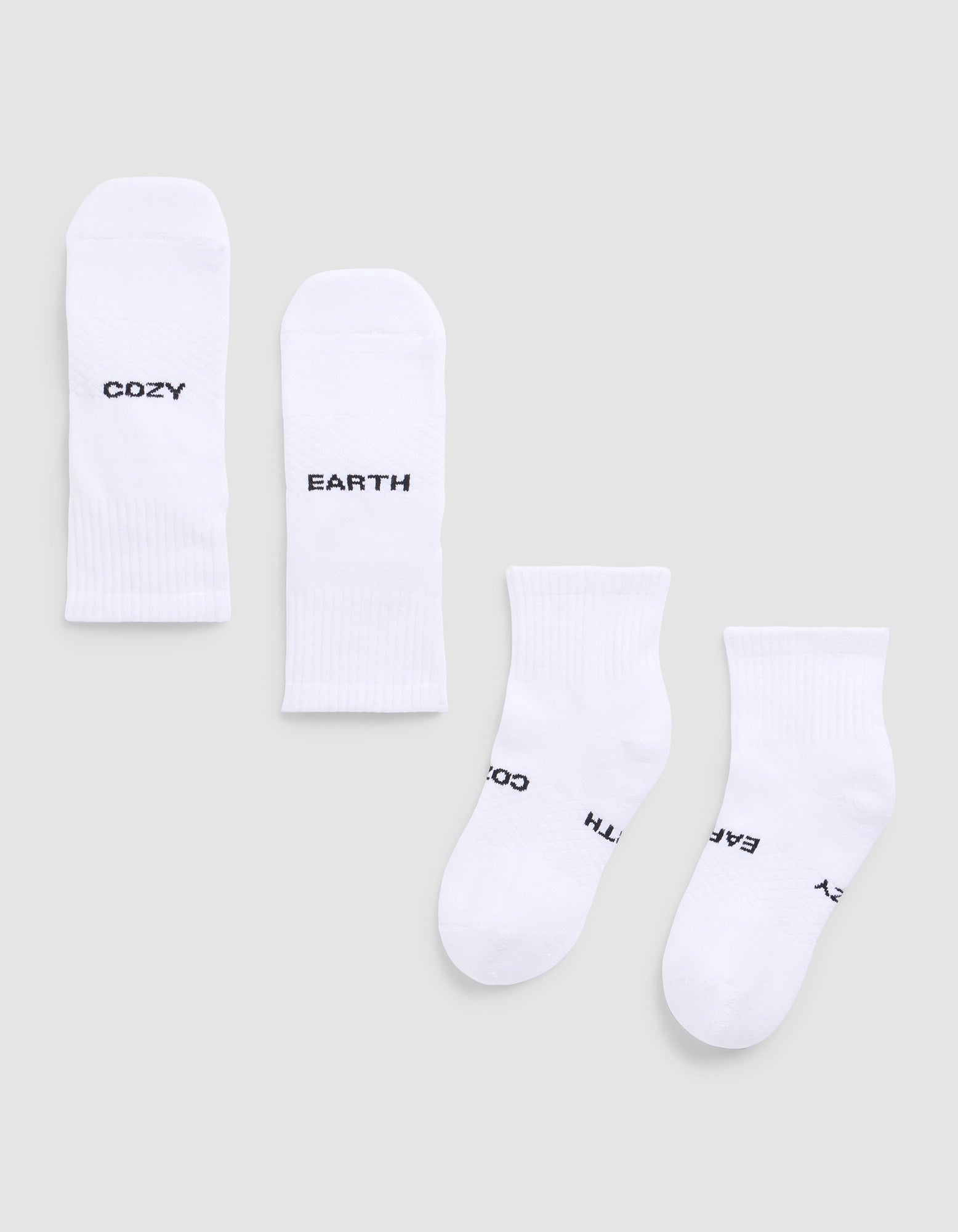 Four white Essential Quarter Socks from Cozy Earth are laid flat on a light gray background. Each features black text: one pair reads “COZY” and “EARTH,” while the other’s text shows parts of “EARTH,” “COZY,”.  