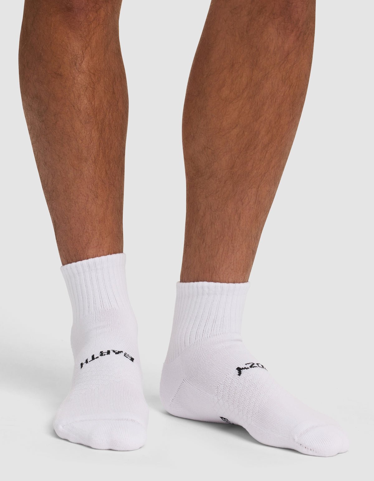 A person wearing Cozy Earth Essential Quarter Socks, featuring black text, stands on a white surface with only their lower legs and feet visible. 