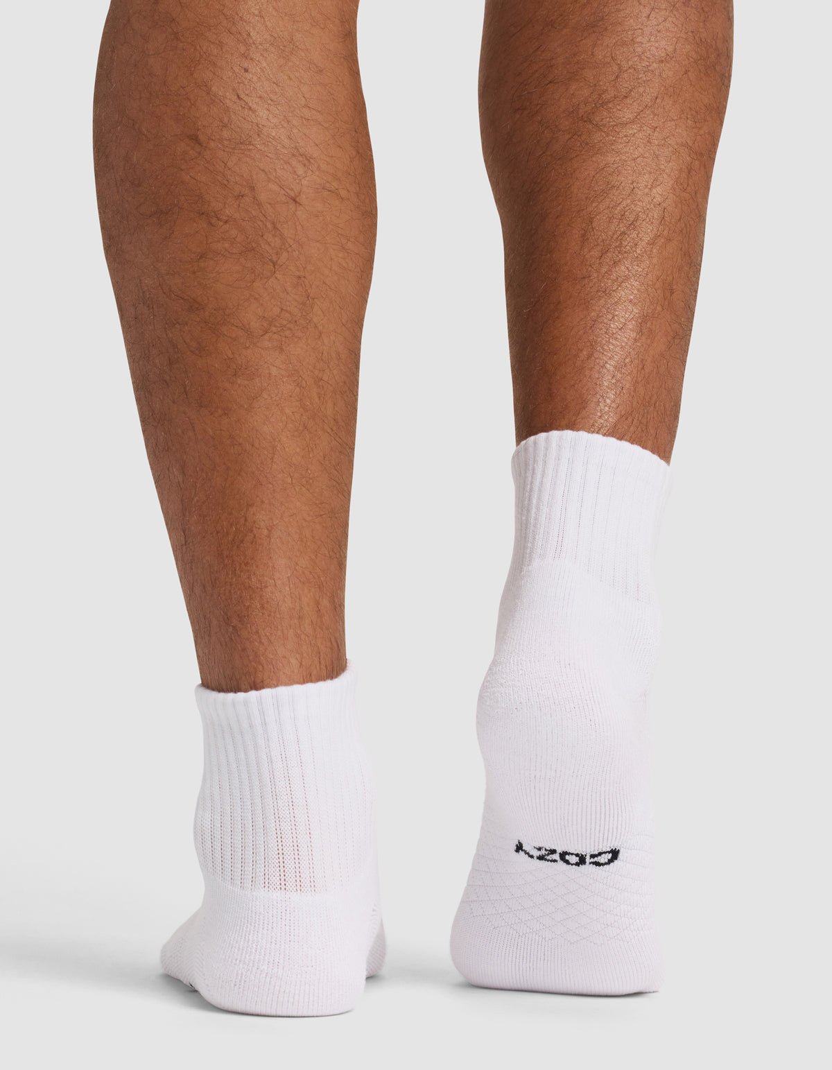 The image shows the back view of a person’s lower legs and feet wearing Cozy Earth Essential Quarter Socks in white. The right sock has "COZY" printed on the heel in black text, set against a plain white background.