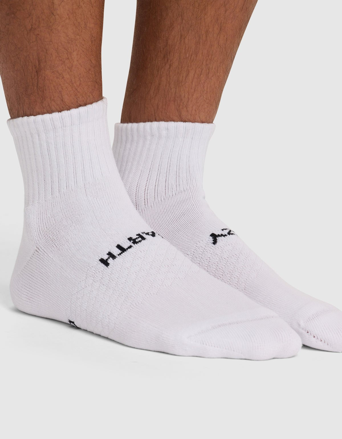 A close-up of a person wearing Cozy Earth Essential Quarter Sock 4-Pack, white ribbed crew socks with black text near the arch, standing on a white surface.