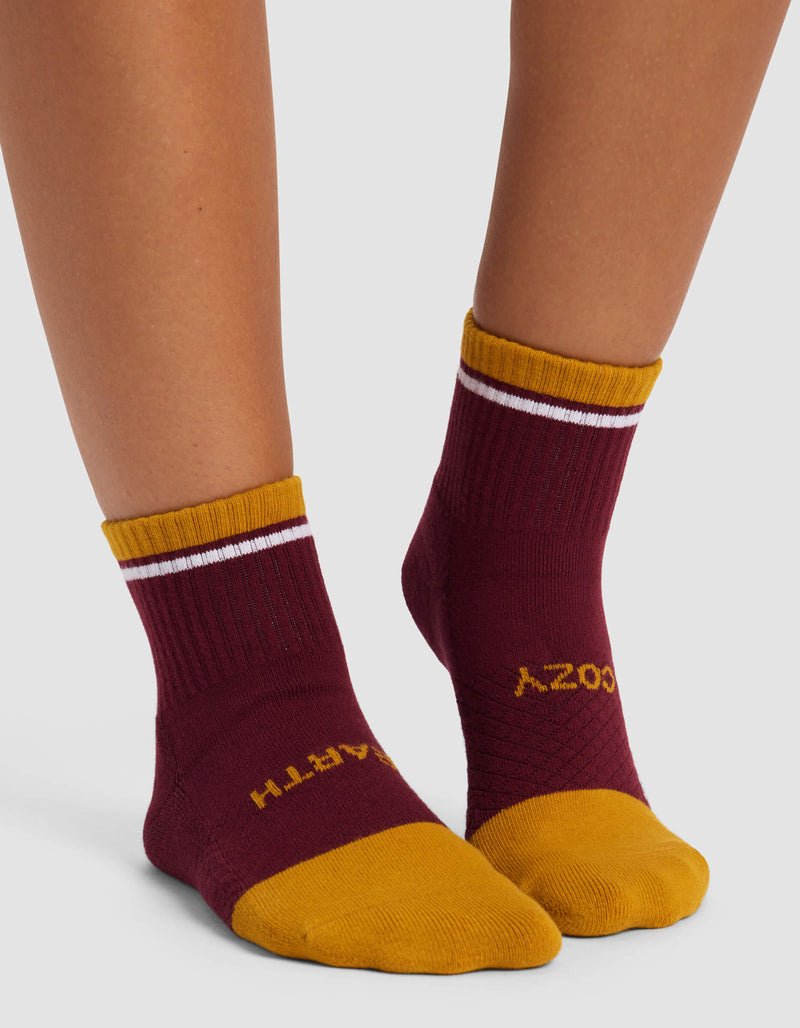 A person wears Cozy Earth Women's Essential Quarter Socks in maroon with gold toes, heels, and cuffs featuring a white stripe; "EARTH" is on the left sock and "COZY" on the right in yellow letters.