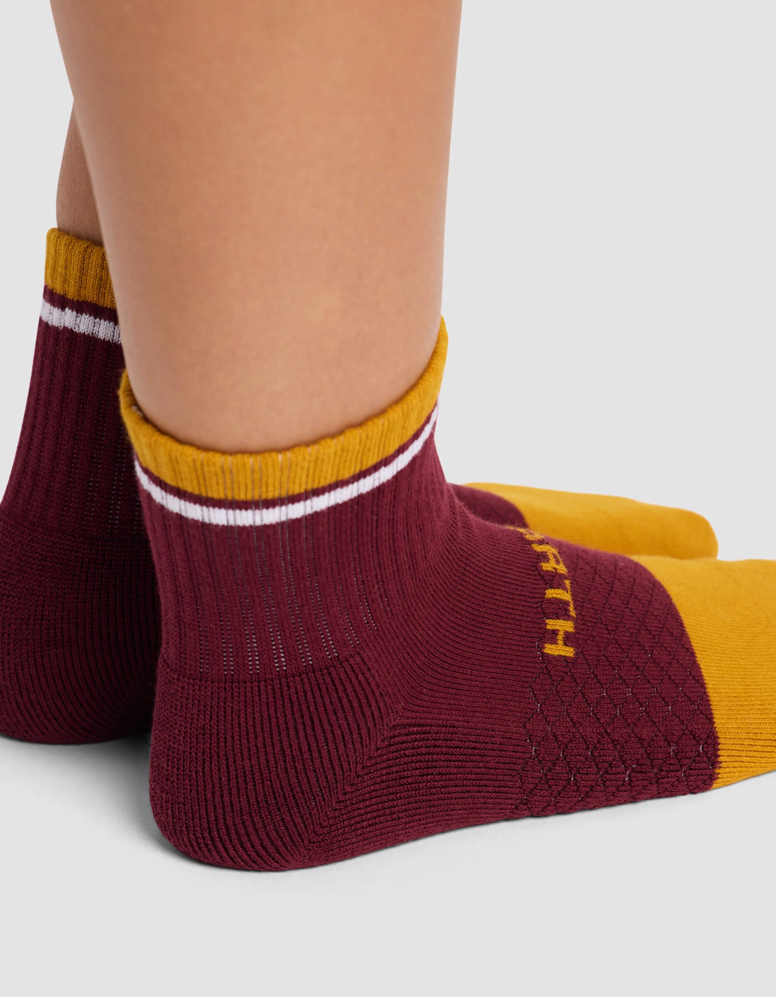 A close-up of two feet wearing Cozy Earth's Women's Essential Quarter Socks in maroon and gold with white cuff stripes, yellow toes, a diamond side pattern, and "EARTH" visible on the socks.