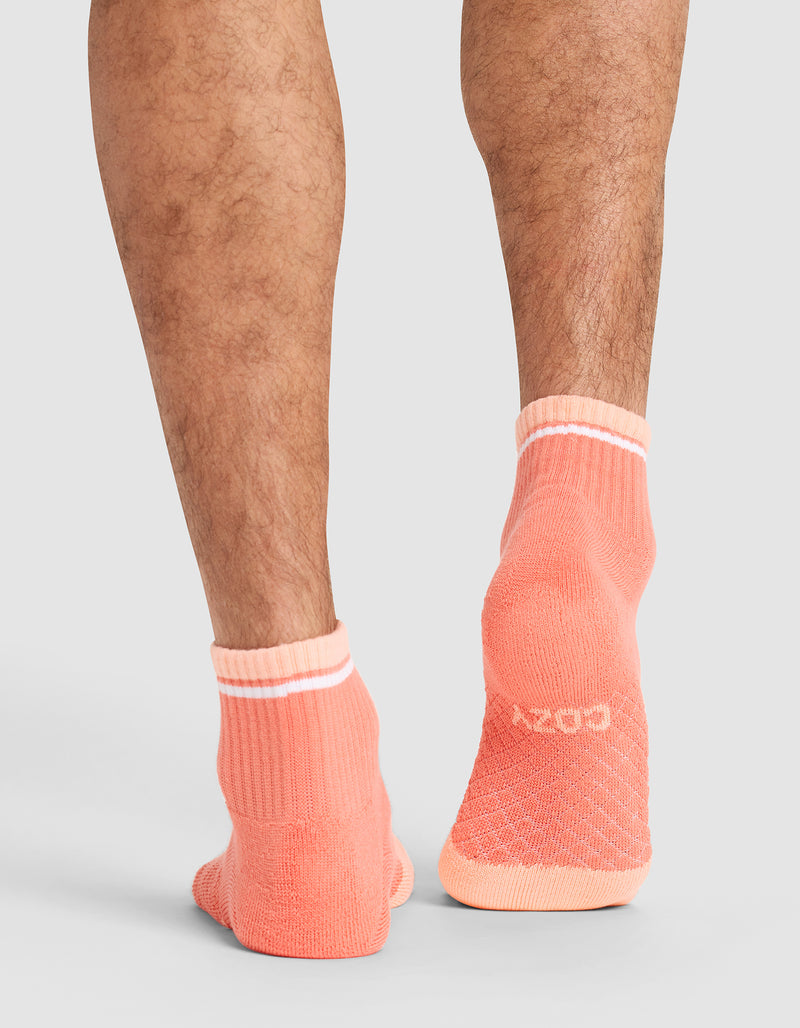 Back view of a person’s lower legs wearing Cozy Earth Essential Quarter Socks in coral with white cuffs and a white logo on the heel, set against a light gray background.