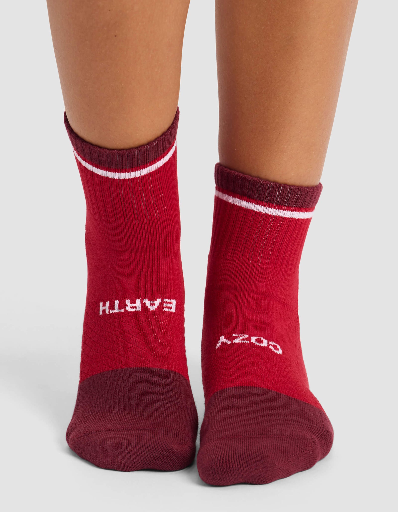 A person wears Cozy Earth's Women's Essential Quarter Socks in red with maroon toes and cuffs, "EARTH" on the left sock and "COZY" on the right, both in white lettering across the top of the feet, standing on a white surface.