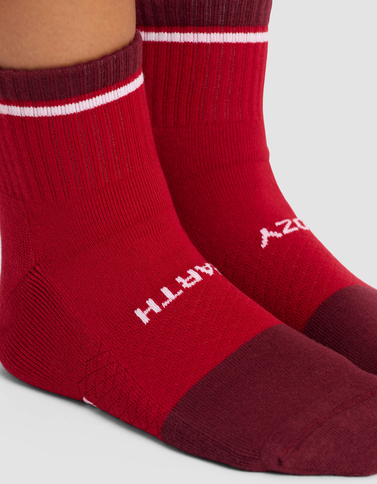 Close-up of two feet wearing Cozy Earth's Women's Essential Quarter Sock in red with a darker maroon toe and heel, a white stripe near the top, and upside-down white "EARTH" text on the side.