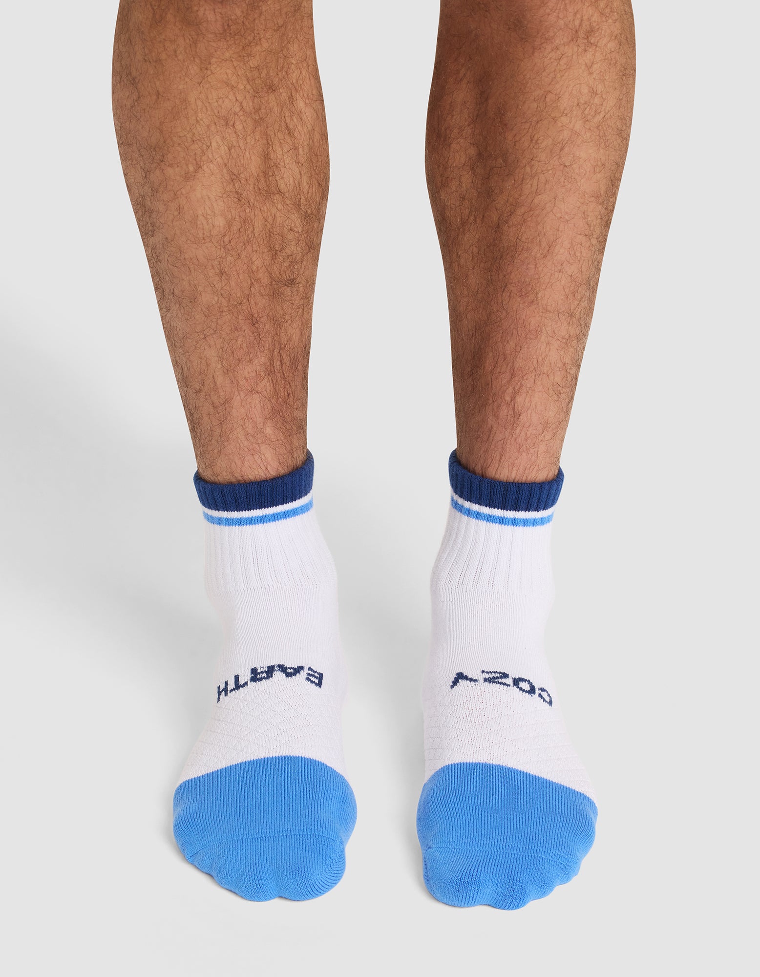 A person stands on a light surface wearing Cozy Earth Essential Quarter Socks in white with blue toes and cuffs and "EARTH" and "COZY" printed in dark blue on top. |Color:Harbor