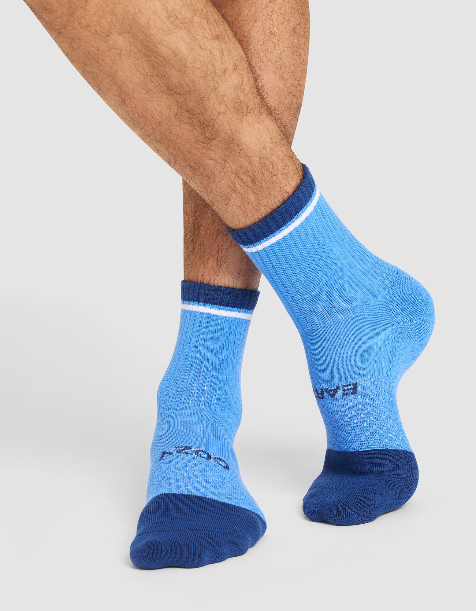 A person with crossed legs wears Cozy Earth Essential Quarter Socks in light blue with dark blue toes, heels, and cuffs, white stripes near the cuffs, and text on top of the foot. The background is light gray. 