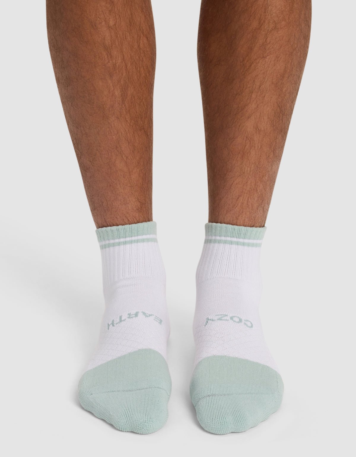 A person wears Cozy Earth Essential Quarter Socks—white with light green toes, cuffs, and "COZY EARTH" text. Only lower legs and feet are visible against a plain background.