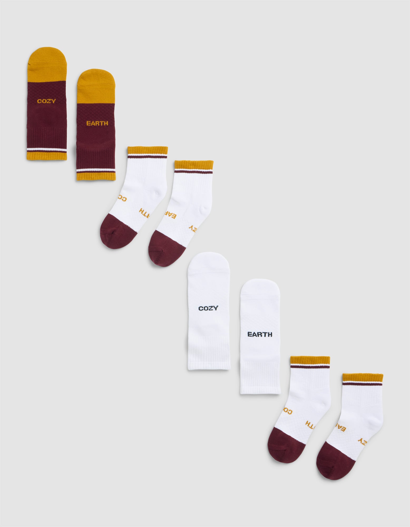 Three pairs from the Cozy Earth Essential Quarter Sock 4-Pack are arranged diagonally on a light gray background, featuring maroon and yellow "COZY EARTH" socks, white socks with gold accents, and plain white "COZY EARTH" socks.|Color:Burgundy
