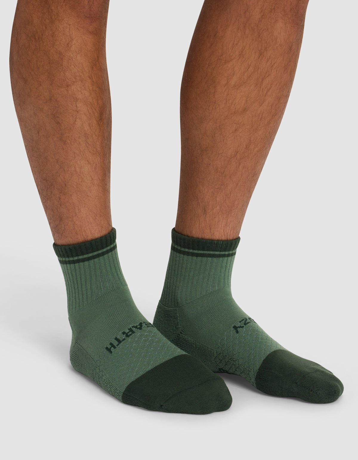 A person wearing Cozy Earth Essential Quarter Socks - Last Chance in dark and light green, featuring a textured pattern, ribbed cuffs, and partially visible text, stands on a white background.|Color:Spruce