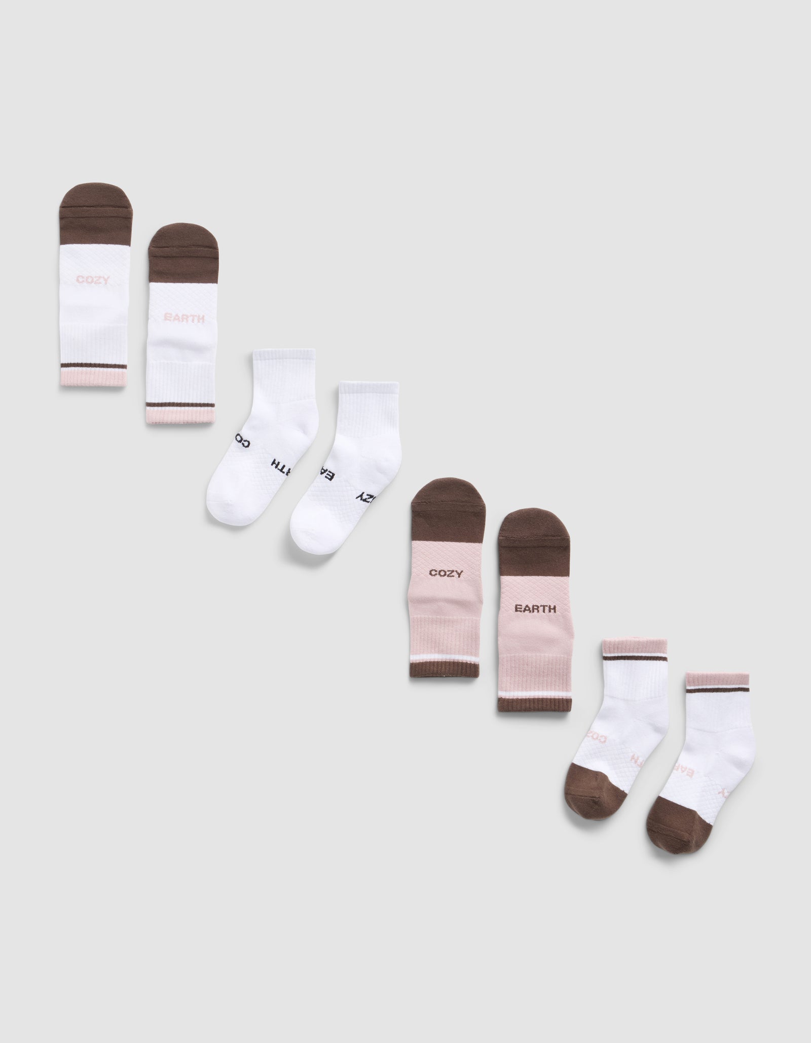 A diagonal row of six pairs of Essential Quarter Sock 4-Packs by Cozy Earth in brown, white, and pink on a light gray background. Some socks feature printed text like "COZY," "EARTH," 