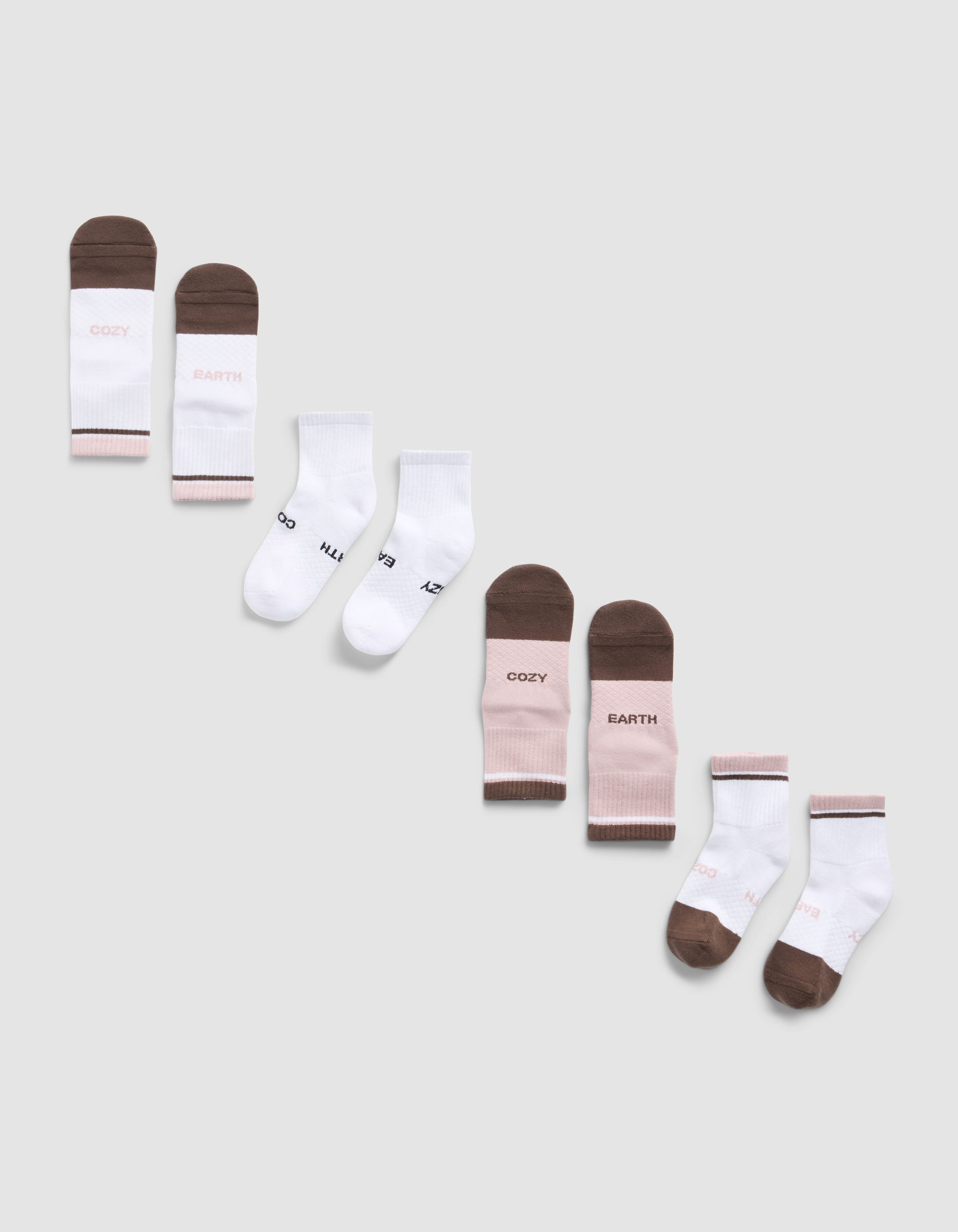 A diagonal row of six pairs of Essential Quarter Sock 4-Packs by Cozy Earth in brown, white, and pink on a light gray background. Some socks feature printed text like "COZY," "EARTH," 