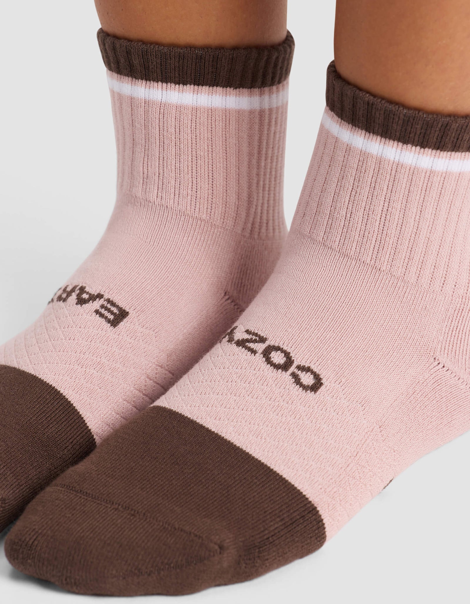 Two feet wear Cozy Earth's Women's Essential Quarter Socks in light pink with brown toes, heels, and cuffs, plus a white stripe near the cuff and black text on top. The background is plain and light-colored.