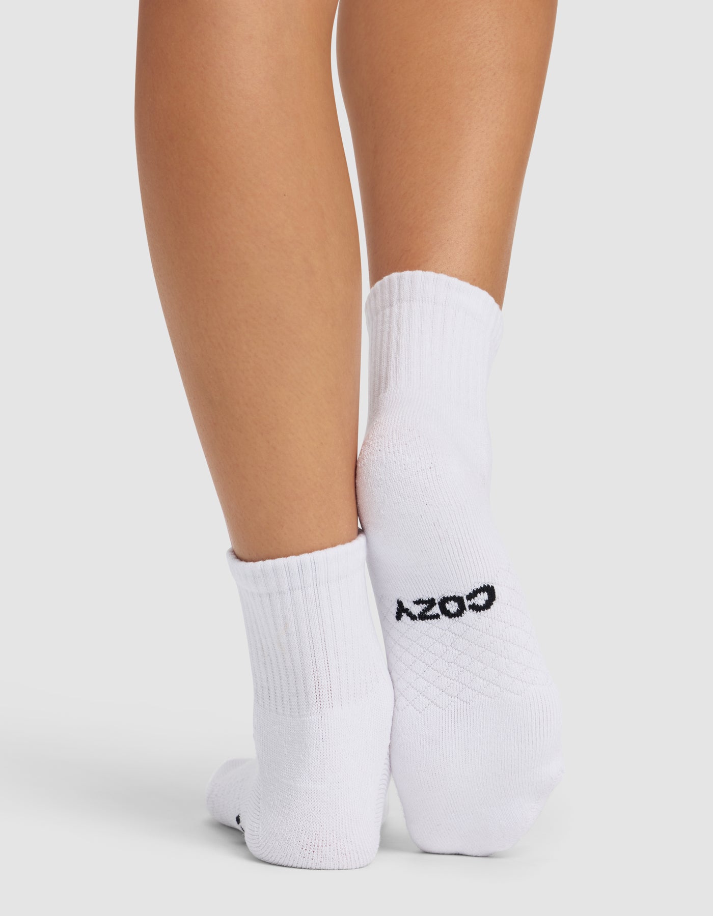 A person stands with feet crossed, wearing Cozy Earth Essential Quarter Socks in white. "COZY" is written in black on the sole of one sock. The background is plain and light-colored.