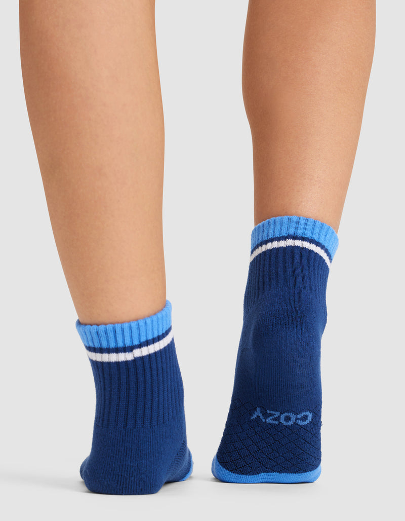 A person wearing Cozy Earth Essential Quarter Socks in blue with light blue trim and a white stripe stands on a white background. The socks feature ribbed cuffs and "cozy" printed on the sole.