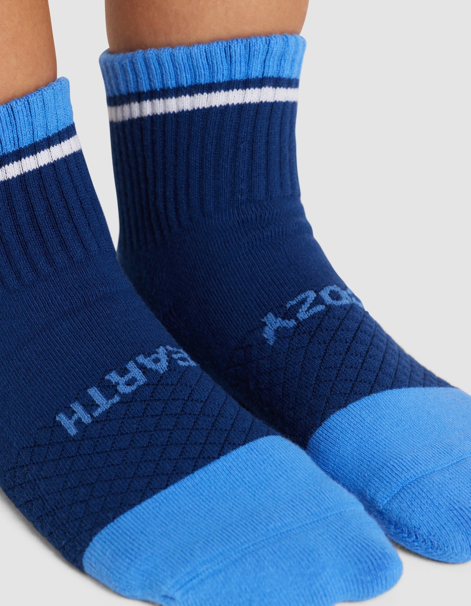 Close-up of feet wearing Cozy Earth Essential Quarter Socks in blue, featuring light blue toes and heels, white and dark blue stripes near the cuff, and partial white lettering on top. 