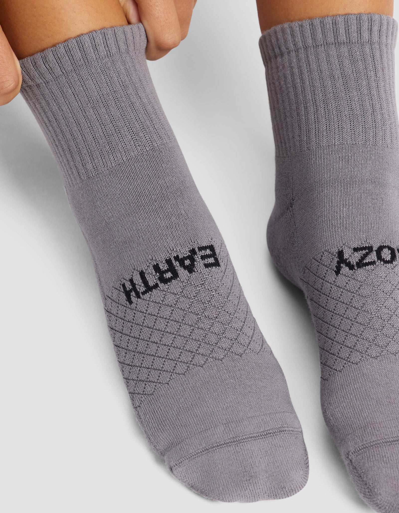 A close-up of two feet in Cozy Earth's Women's Essential Quarter Socks, light grey with ribbed cuffs. "EARTH" is written on the left sock and "COZY" on the right in black text across the top.