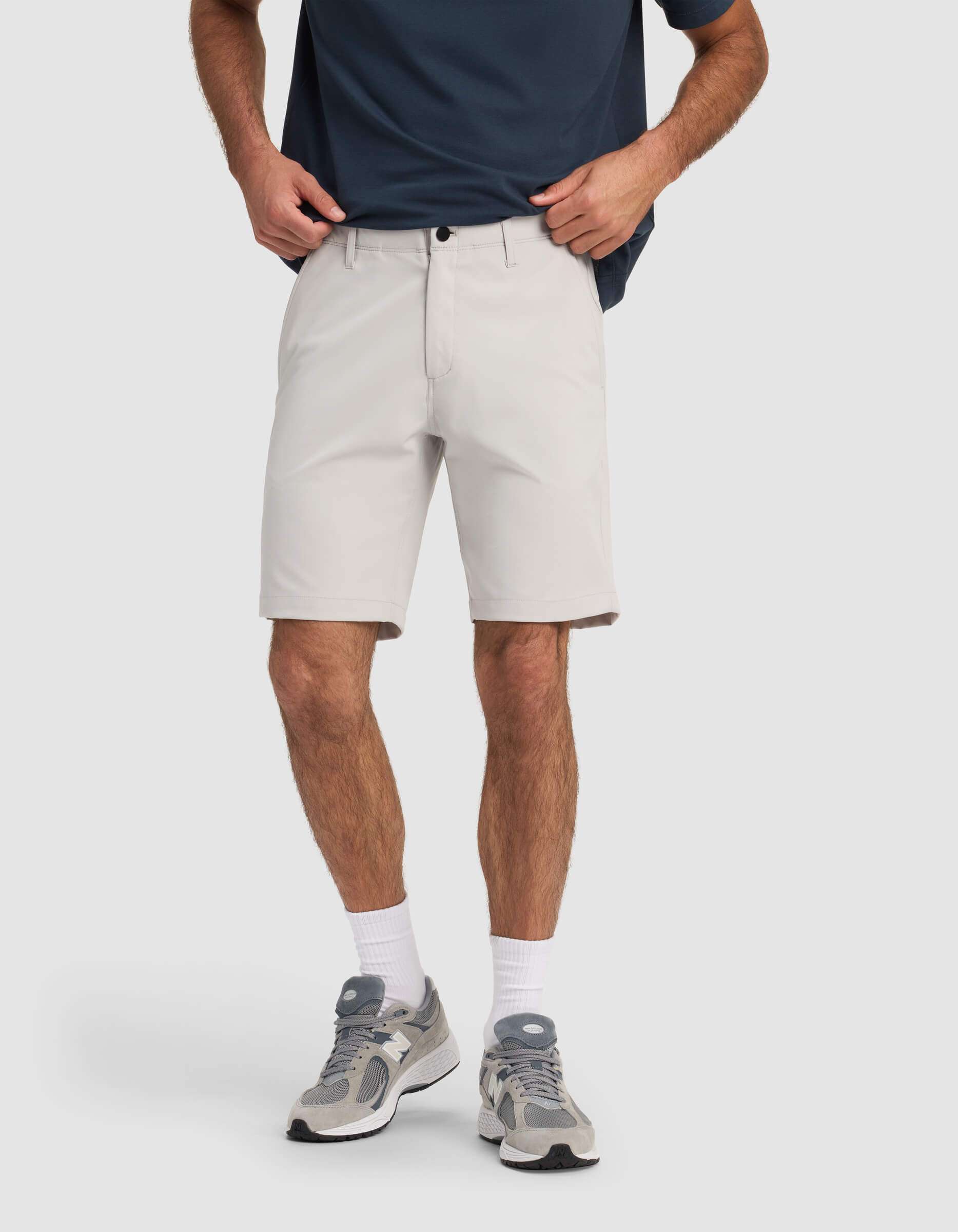 A person models Cozy Earth's Men's Everywhere Short in light gray with a navy shirt, white socks, and gray sneakers. They stand against a plain white background with hands in pockets; their face is not visible.