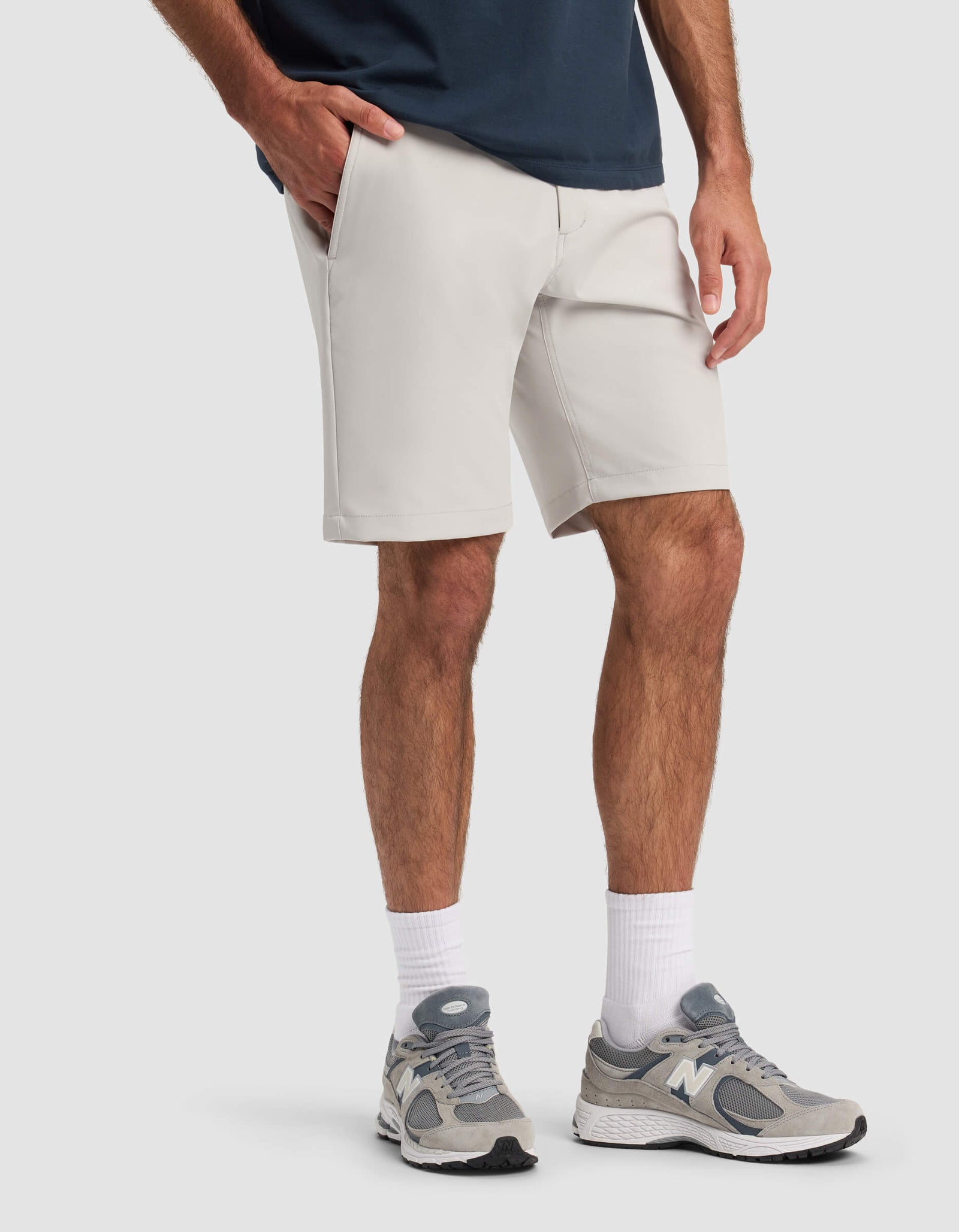 A person wearing Cozy Earth Men's Everywhere Short in light gray, a navy shirt, white crew socks, and gray New Balance sneakers stands with one hand in their pocket against a plain light background.