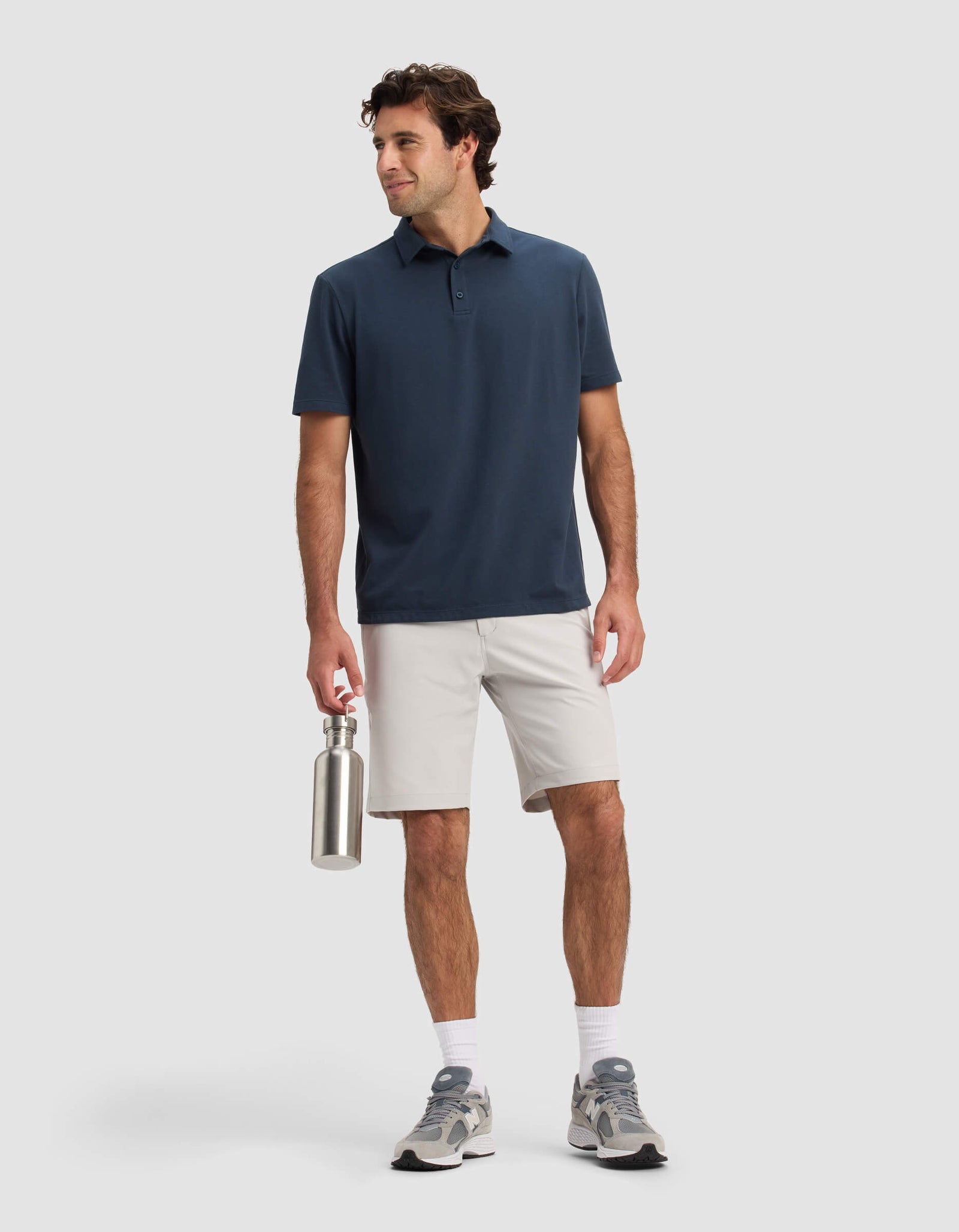A man stands smiling against a plain background, wearing the Cozy Earth Men's Everywhere Short in light gray, paired with a navy polo, white socks, gray sneakers, and holding a metal water bottle.