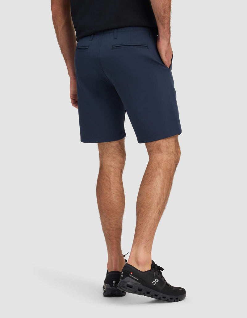 A person wearing Cozy Earth's Men's Everywhere Short in dark blue, with a black shirt and black athletic shoes, stands facing away from the camera with their left hand in their pocket against a plain light background.