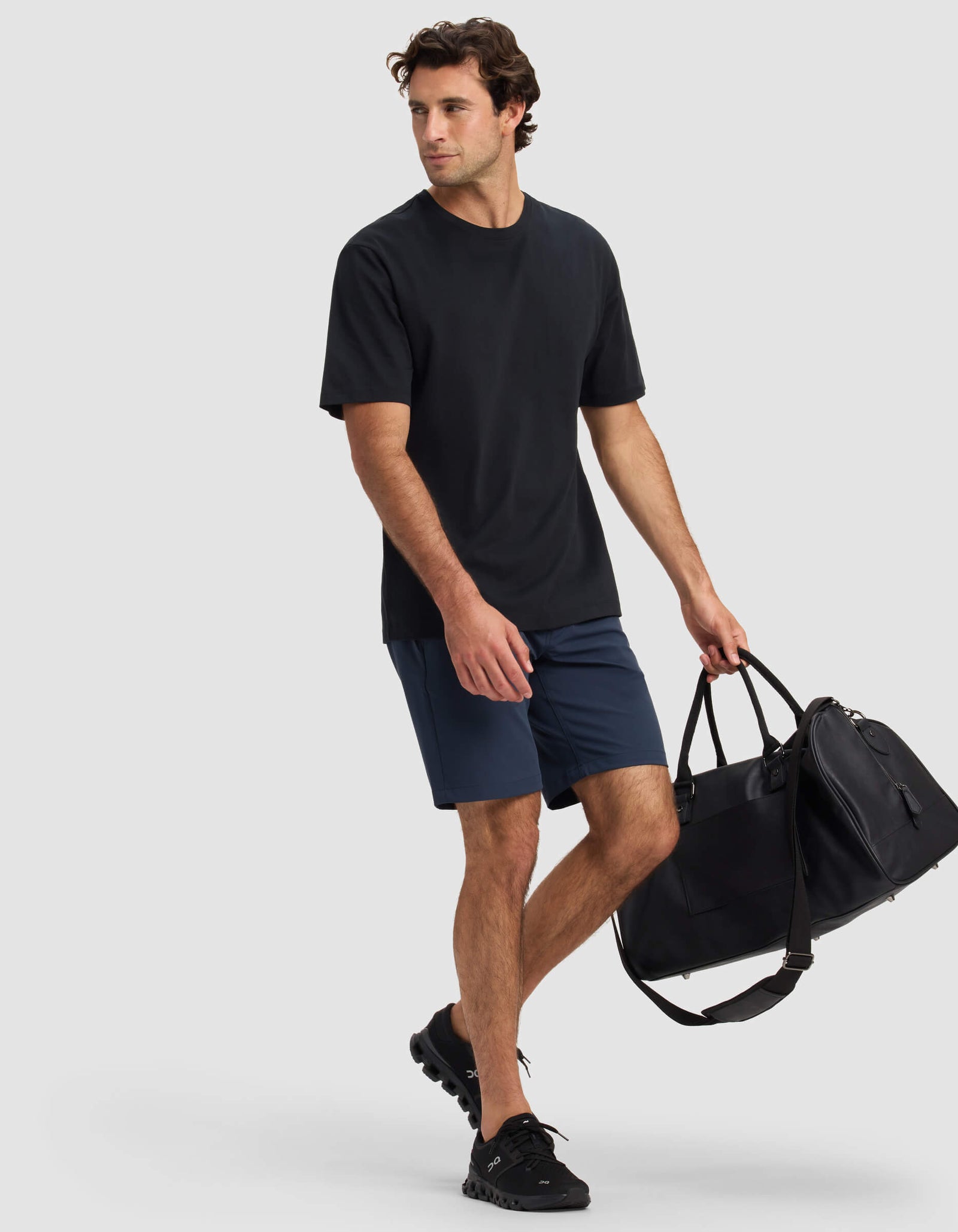 A man wearing Cozy Earth's Men's Everywhere Short in navy and a black t-shirt walks with a large black duffel bag with a shoulder strap and black sneakers against a plain light background.
