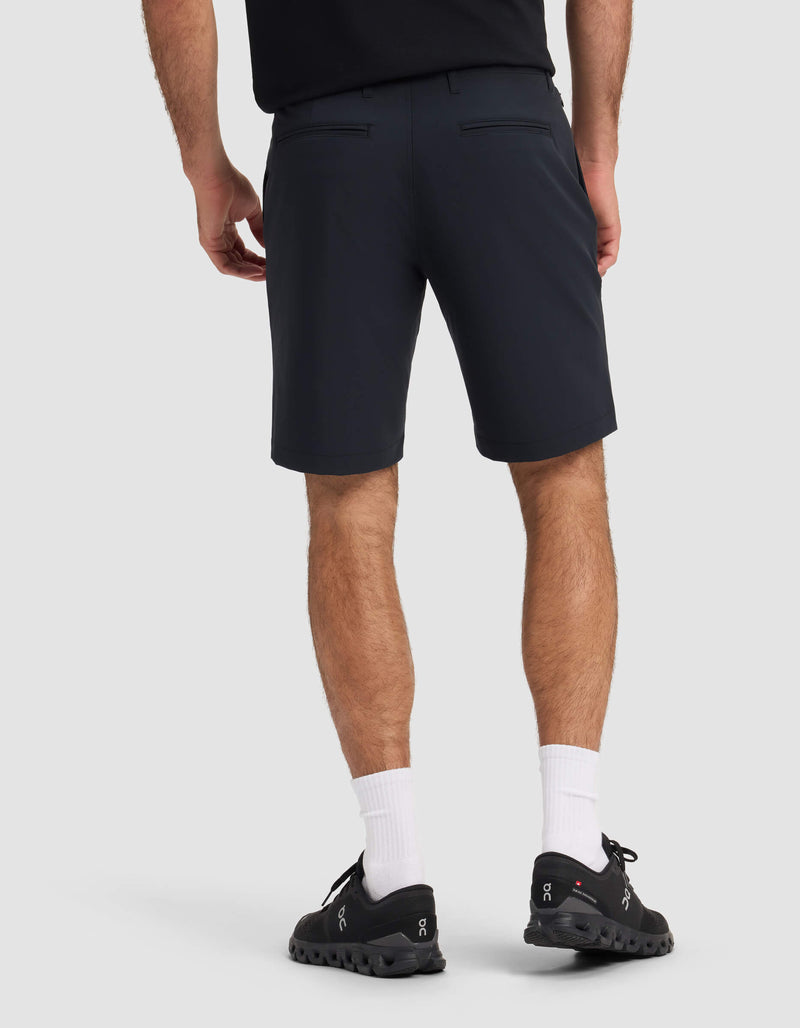 A person is standing facing away, wearing a black shirt, Cozy Earth Men's Everywhere Short in dark navy, white crew socks, and black athletic shoes against a plain light background.