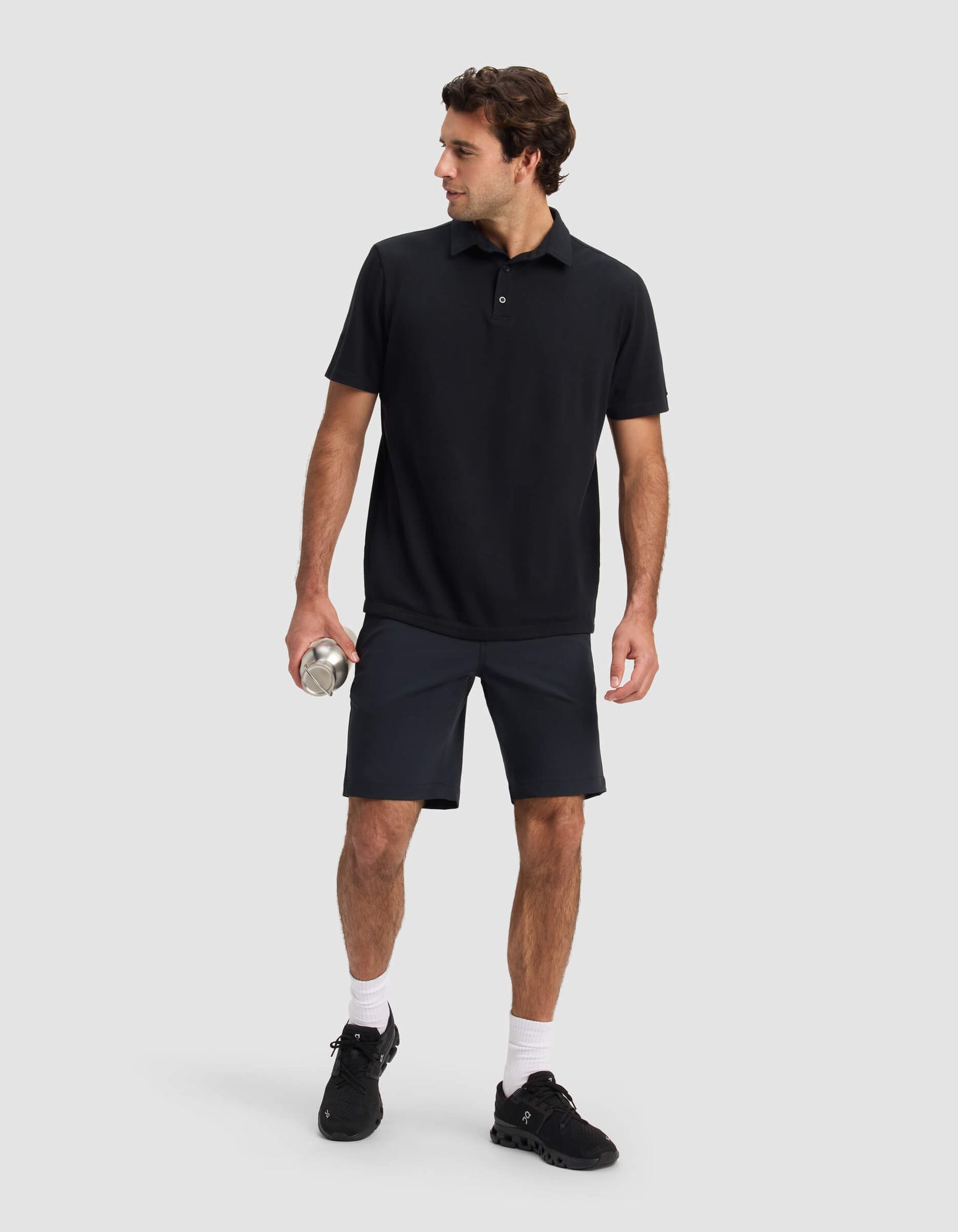A man stands against a light gray background, wearing Cozy Earth's Men's Everywhere Short in black, paired with a black polo shirt, black sneakers, and white socks. He holds a silver water bottle and looks to his left.