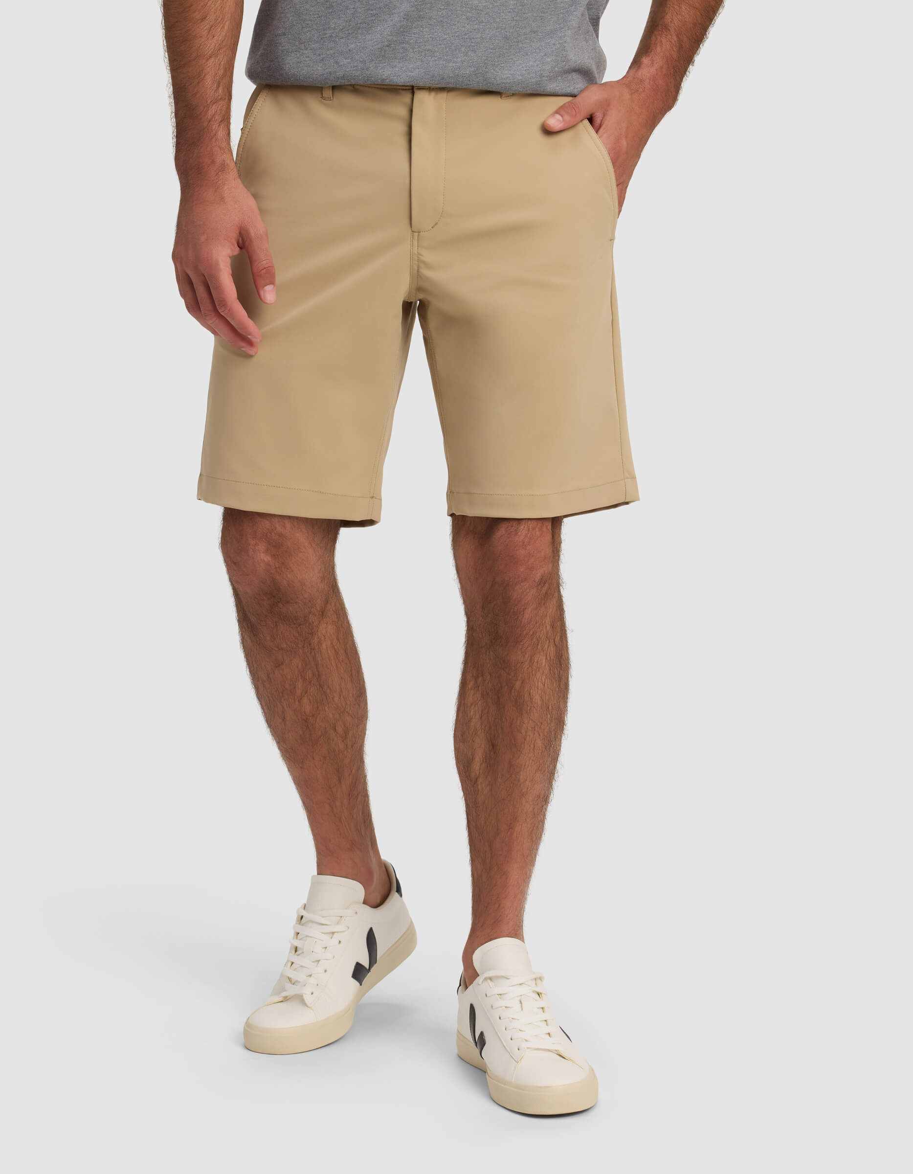 A man wearing Cozy Earth Men's Everywhere Short in beige, a gray t-shirt, and white sneakers with black accents stands with one hand in his pocket against a plain background.|Color:Sandalwood