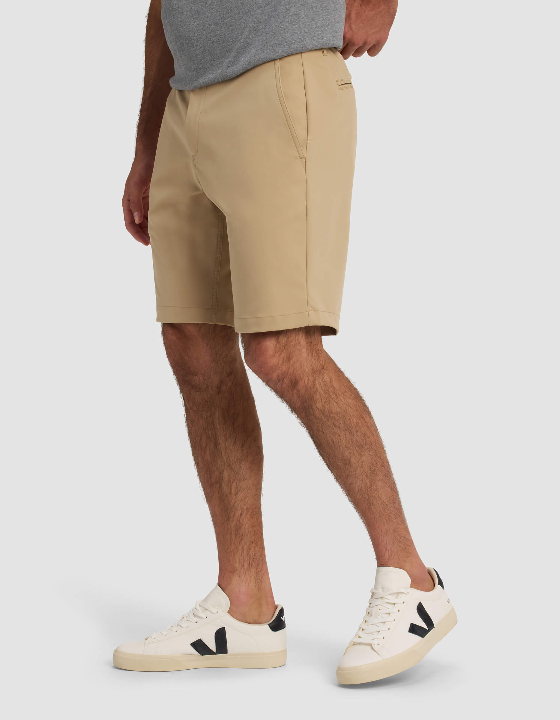 A person wears Cozy Earth's Men's Everywhere Short in beige, a gray t-shirt, and white sneakers with black detail while standing against a plain light background.|Color:Sandalwood