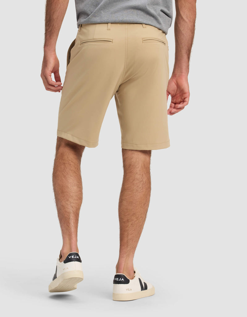 A man stands with his back to the camera against a plain light background, wearing Cozy Earth Men's Everywhere Short in tan, a gray shirt, and white Veja sneakers.