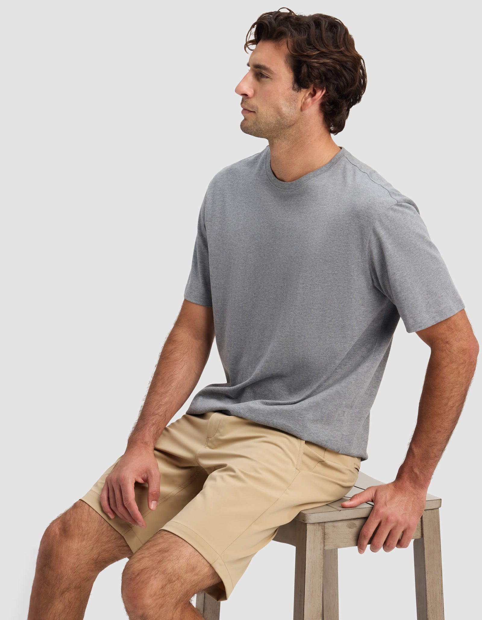 A man with wavy brown hair, wearing a gray t-shirt and Cozy Earth Men's Everywhere Short in beige, sits on a wooden stool against a plain light background, looking to his left.