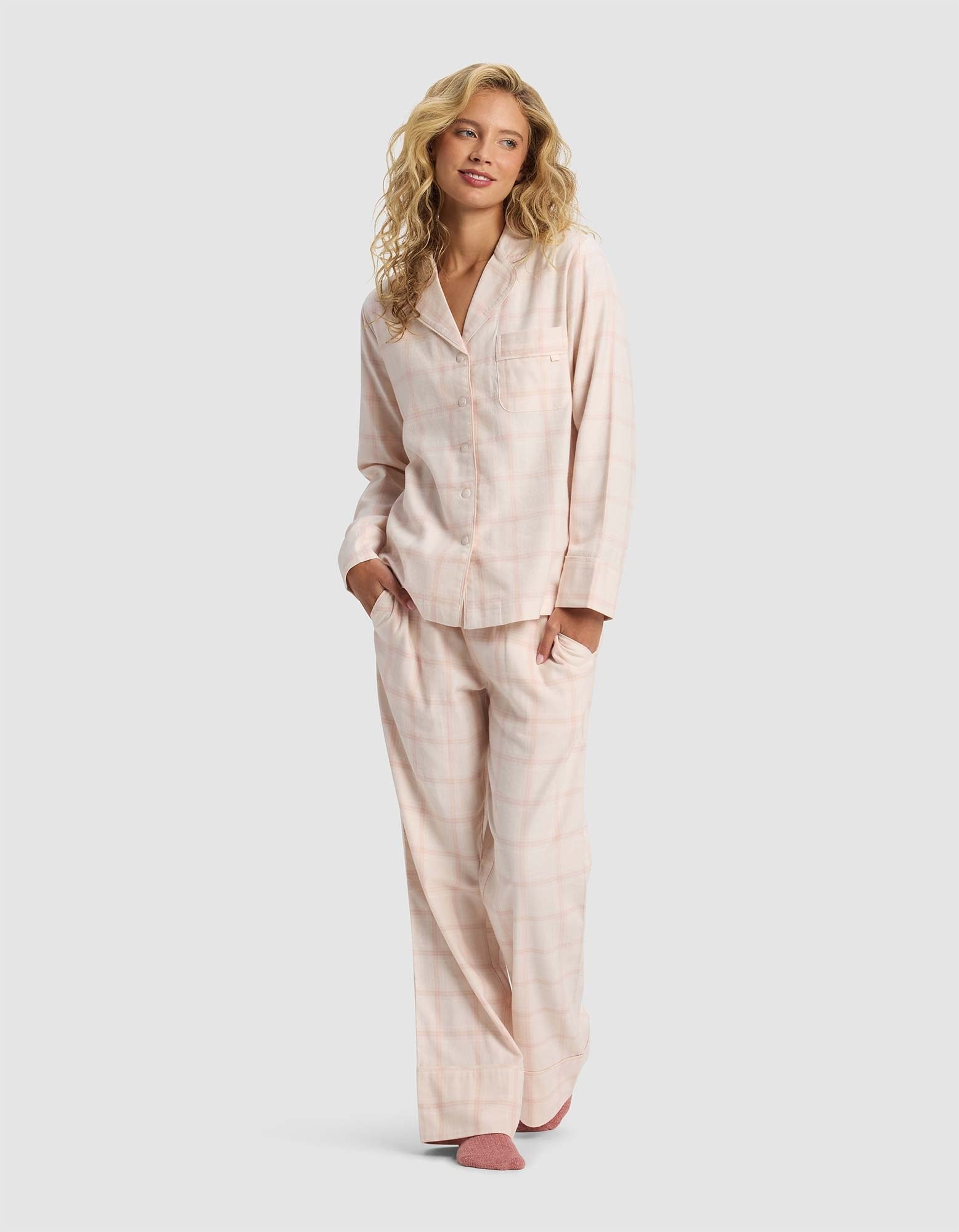 A woman with long blonde hair smiles and stands with her hands in her pockets, modeling the Cozy Earth Women's Farmhouse Long Sleeve Pajama Set in light pink plaid against a plain gray background.|Color:Quartz Plaid