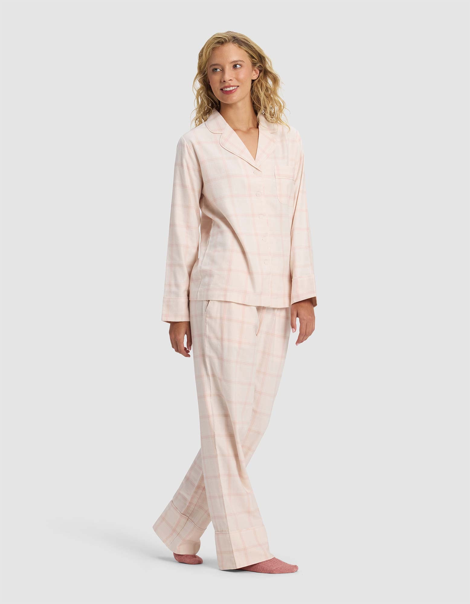 A woman smiles wearing Cozy Earth's Women's Farmhouse Long Sleeve Pajama Set—light pink plaid top and pants with pink socks—standing against a plain light gray background.