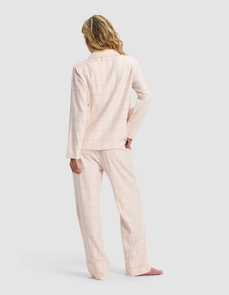 A woman with wavy blonde hair stands with her back to the camera, wearing Cozy Earth's Women's Farmhouse Long Sleeve Pajama Set in light pink plaid and pink slippers against a plain light gray background.