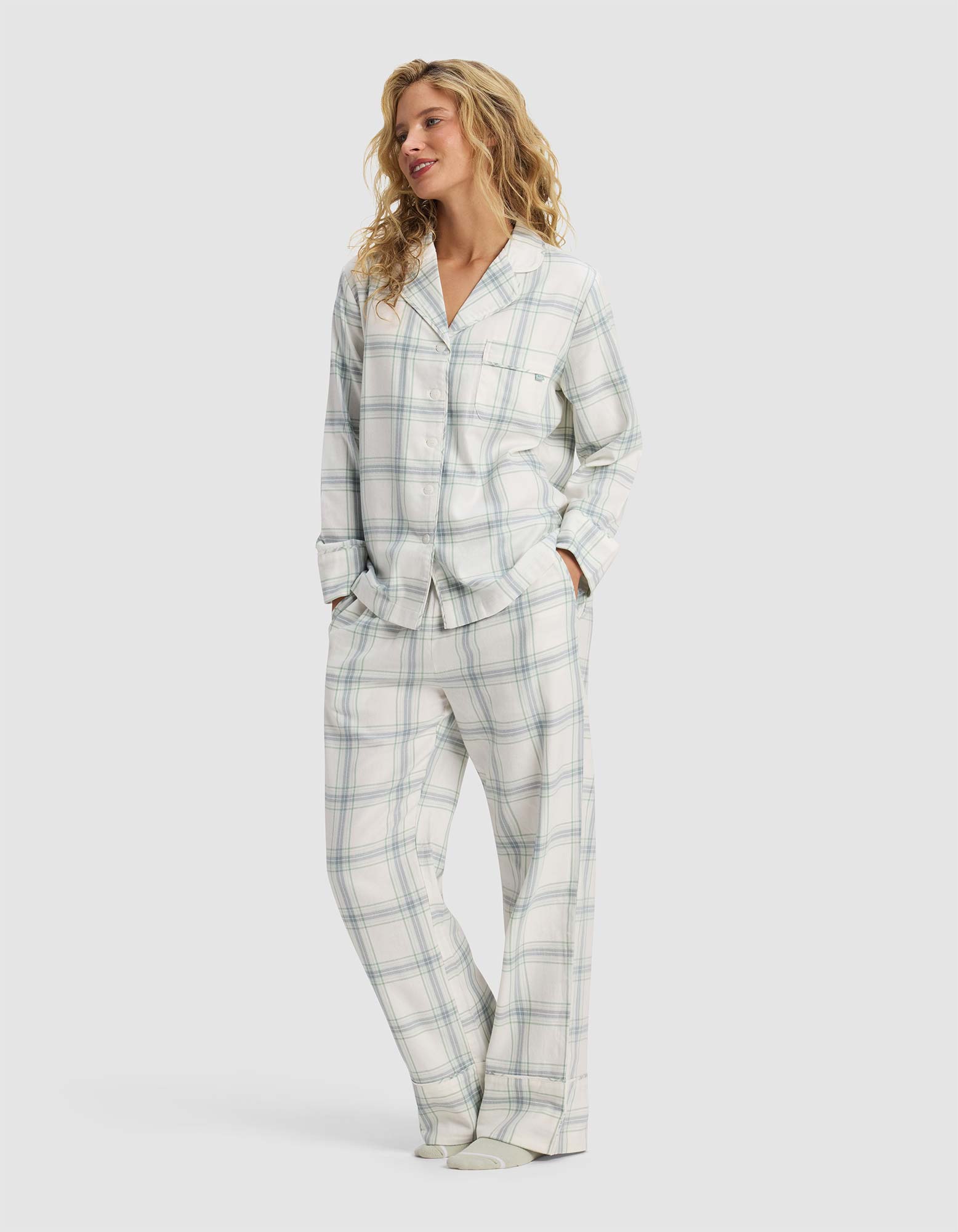 A woman with wavy blonde hair smiles and looks to the side, hands in her pockets, wearing the Cozy Earth Women's Farmhouse Long Sleeve Pajama Set—a loose-fitting, button-up plaid ensemble in light colors.