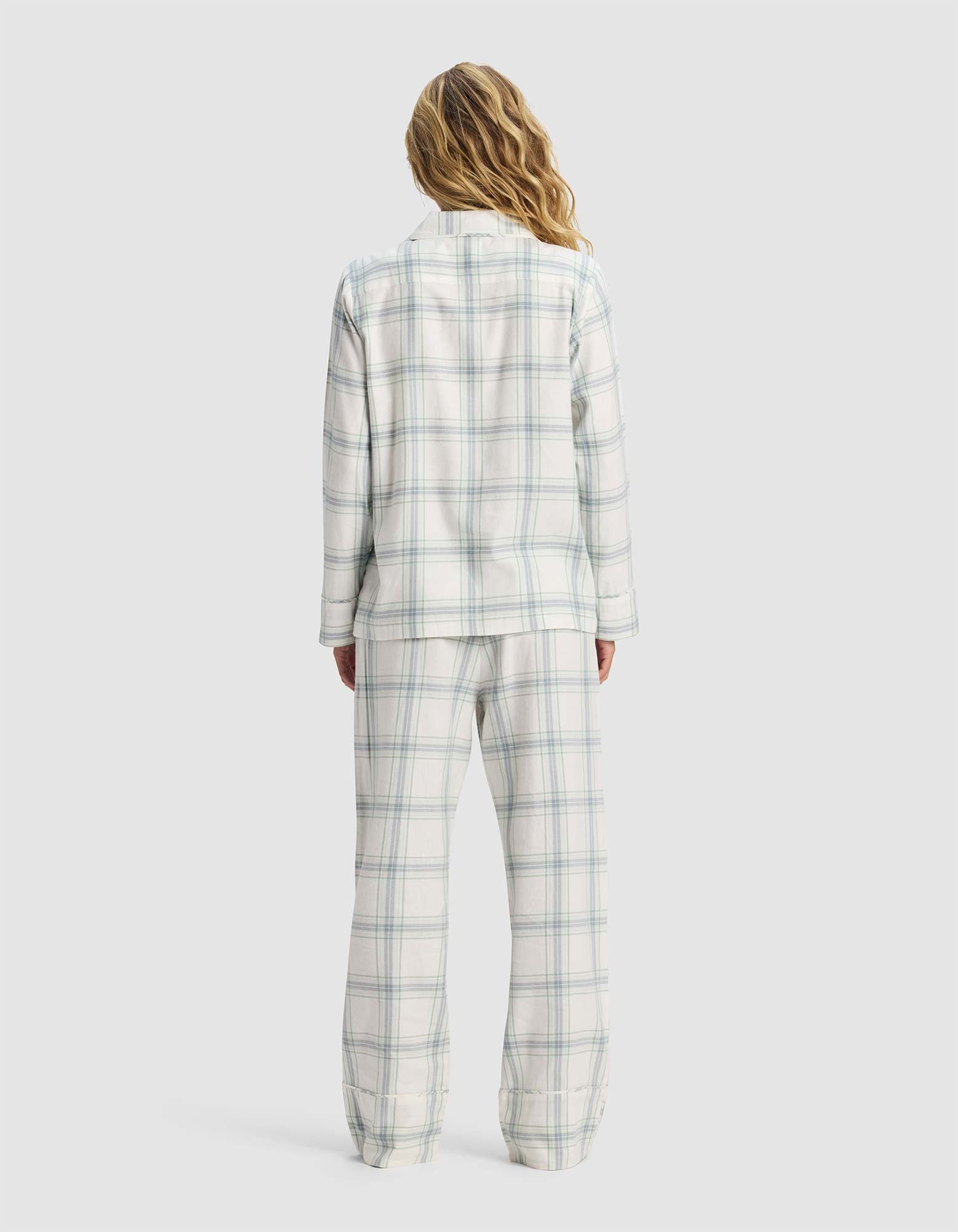 A woman with long, wavy hair stands with her back to the camera, wearing the Cozy Earth Women's Farmhouse Long Sleeve Pajama Set in white and light blue plaid against a plain light background.