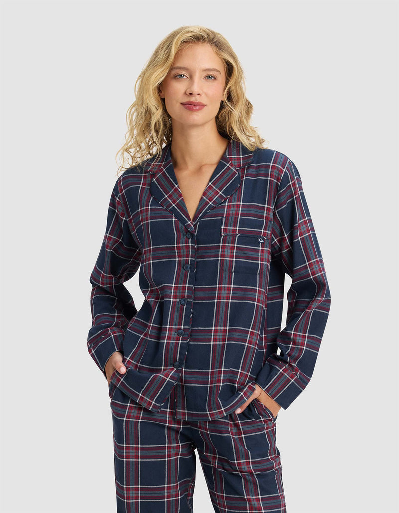 A woman with wavy blonde hair models the Cozy Earth Women's Farmhouse Long Sleeve Pajama Top in a navy, red, and white plaid pattern, standing with hands in her pockets and smiling against a plain light gray background.
