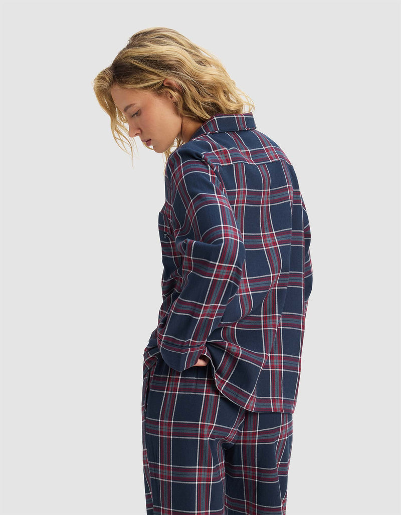 A woman with blond hair stands in profile with hands in pockets, wearing the Cozy Earth Women's Farmhouse Long Sleeve Pajama Top in navy blue and red plaid, paired with matching pants against a plain light gray background.