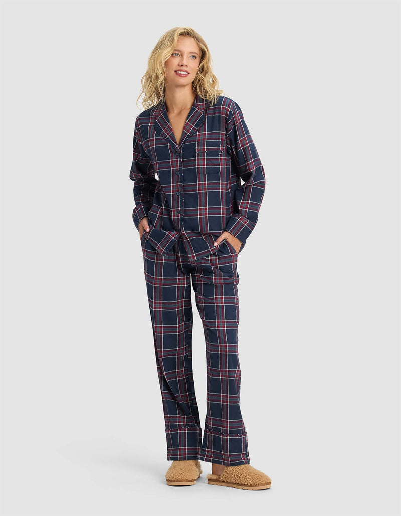 A person stands smiling, wearing the Cozy Earth Women's Farmhouse Long Sleeve Pajama Top in navy blue with red and white plaid, paired with loose pajama pants and tan fuzzy slippers. The top features buttons and pockets.