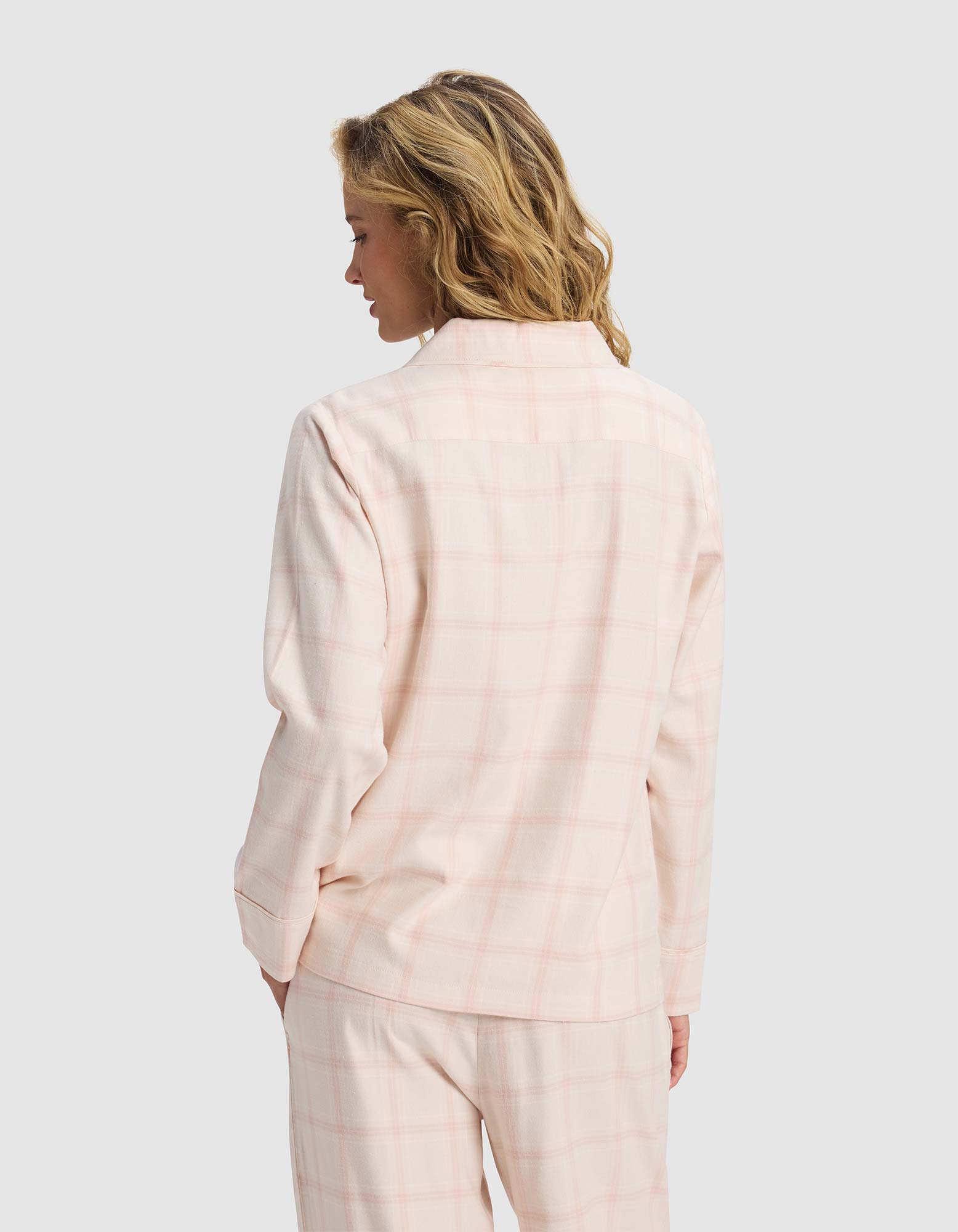 A woman with wavy blonde hair is seen from the back, wearing the Cozy Earth Women's Farmhouse Long Sleeve Pajama Top in pale pink plaid, paired with matching pants, against a plain light gray background.
