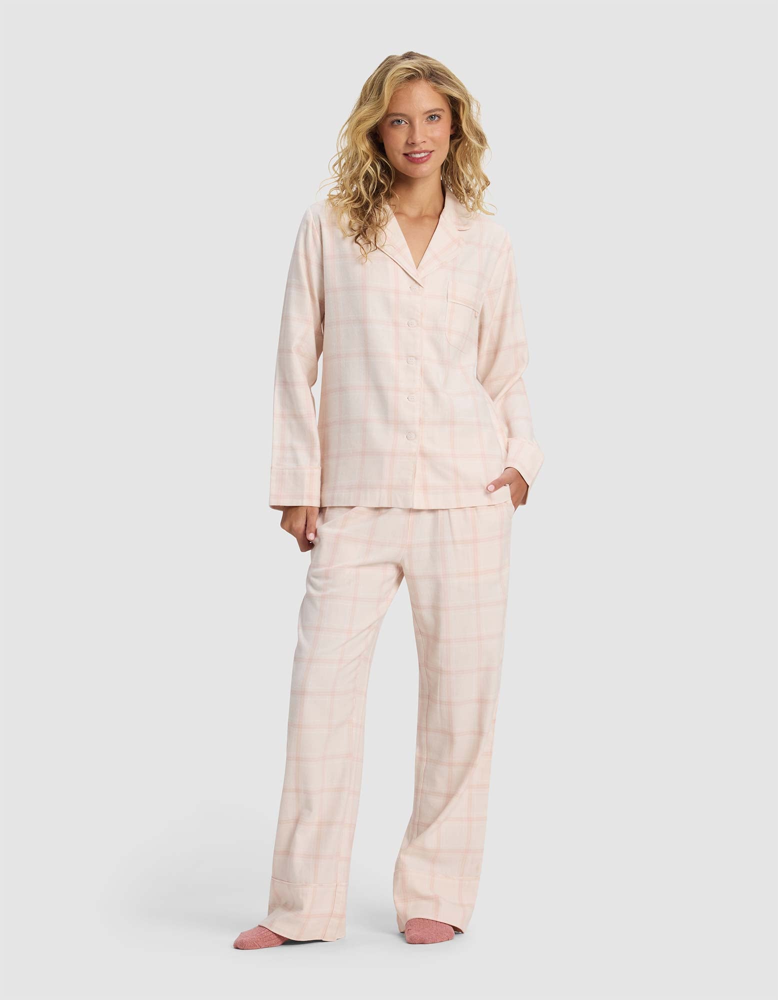 A woman with long blonde hair wears the Cozy Earth Women's Farmhouse Long Sleeve Pajama Top, styled in light pink plaid, paired with pink slippers. She stands against a plain light gray background, smiling slightly with one hand in her pocket.