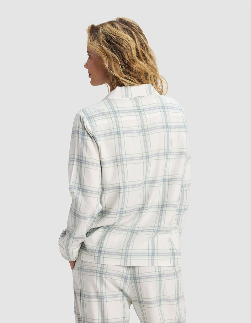A woman with wavy blonde hair stands with her back to the camera, wearing Cozy Earth’s Women's Farmhouse Long Sleeve Pajama Top in white with light blue and green plaid, set against a plain light grey background.