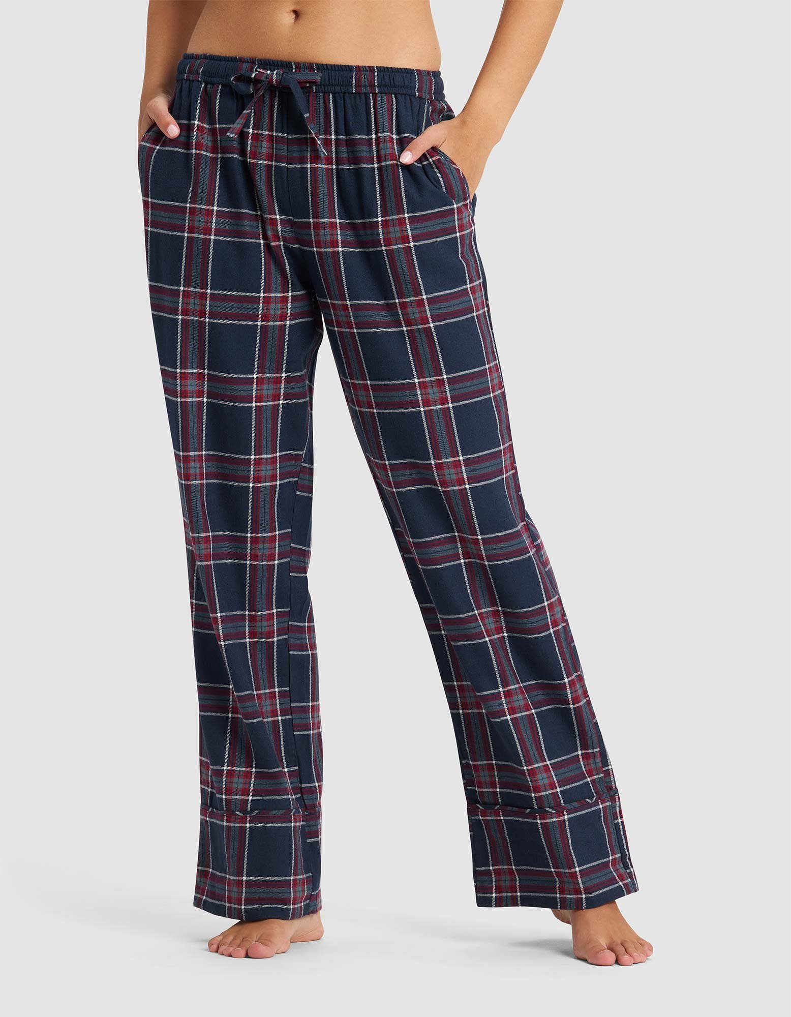 A person stands barefoot with hands in pockets, wearing Cozy Earth Women's Farmhouse Pajama Pant in a navy blue, red, and white plaid pattern. Their upper body is partially out of frame against a plain background.|Color:Eclipse Plaid