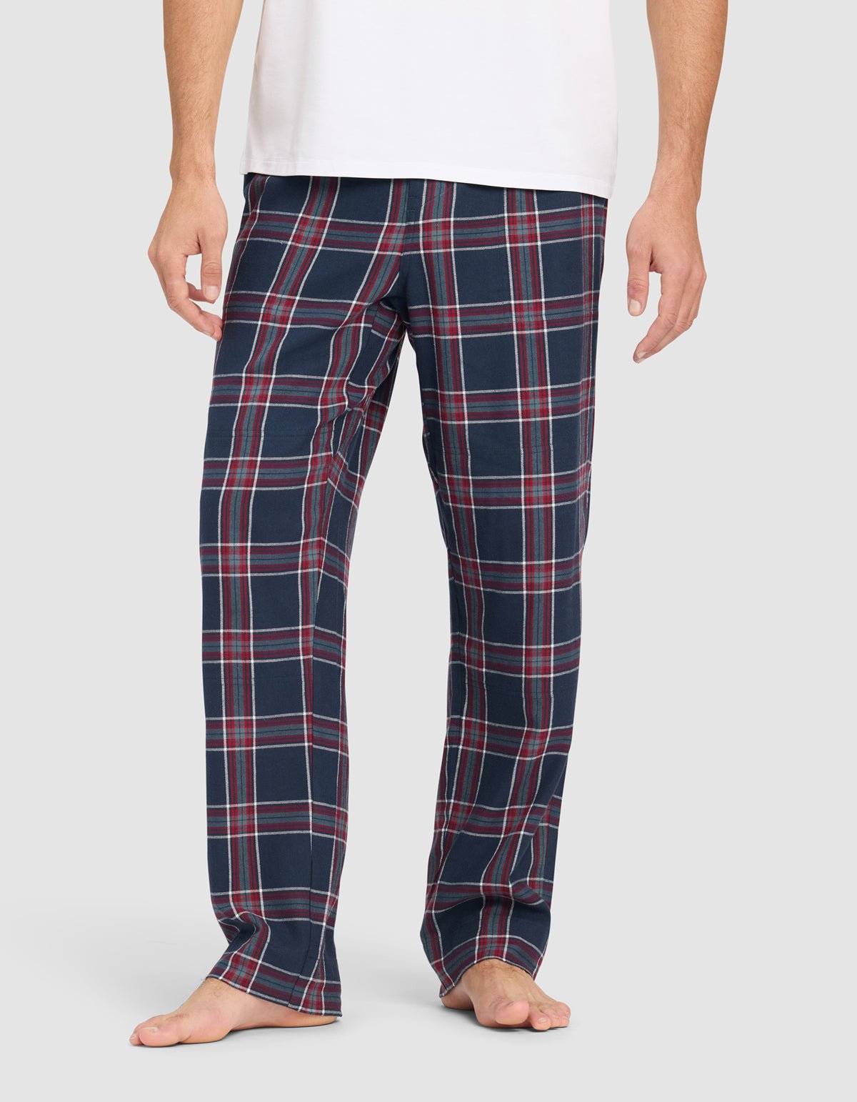 A person wears Cozy Earth Men's Farmhouse Pajama Pant in navy blue plaid with red and white stripes, barefoot against a plain background. The upper body is partially visible, wearing a white shirt.